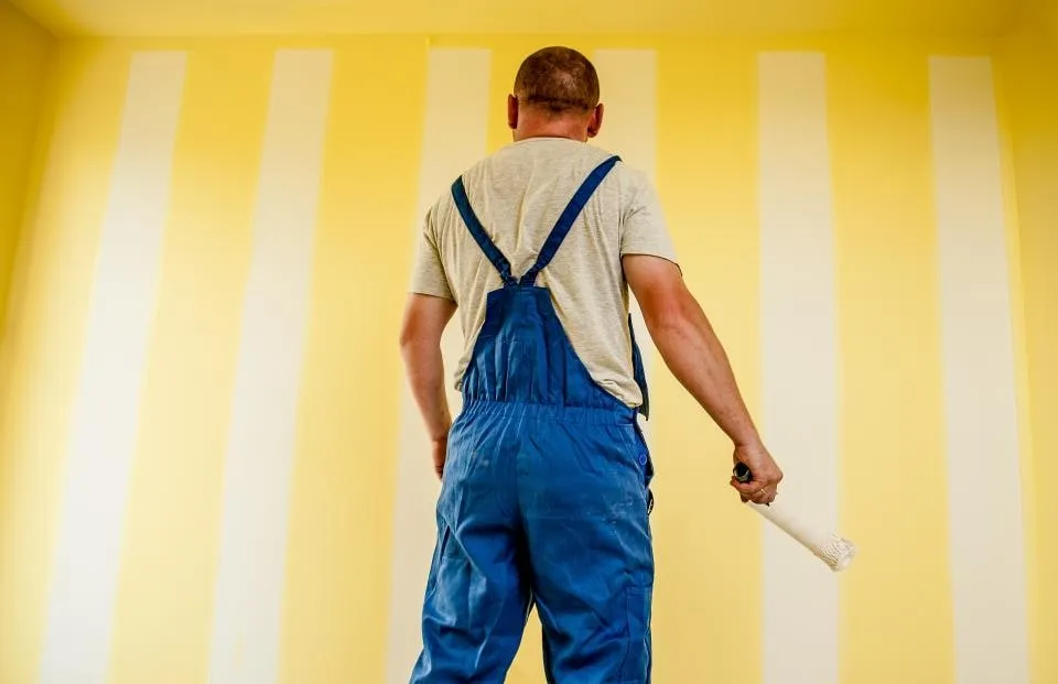 A painter in blue overalls holding a roller, seen from behind, facing a wall of yellow and white stripes